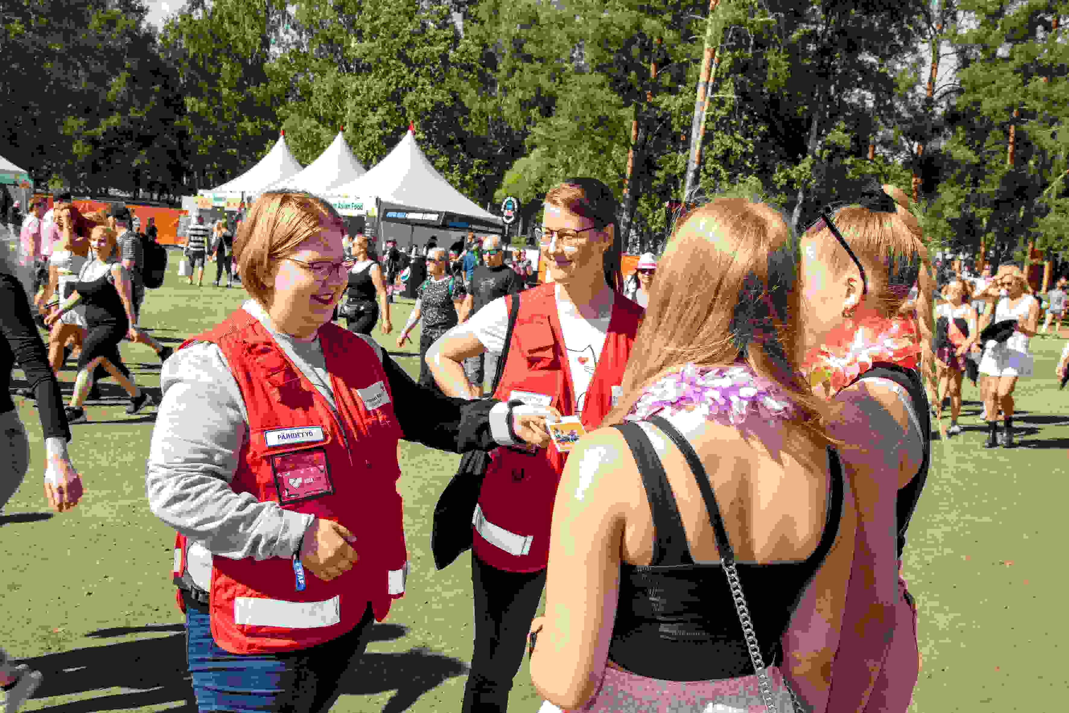 Röda Korsets frivilliga ger två personer en broschyr. I bakgrunden syns en solig festivalmiljö.