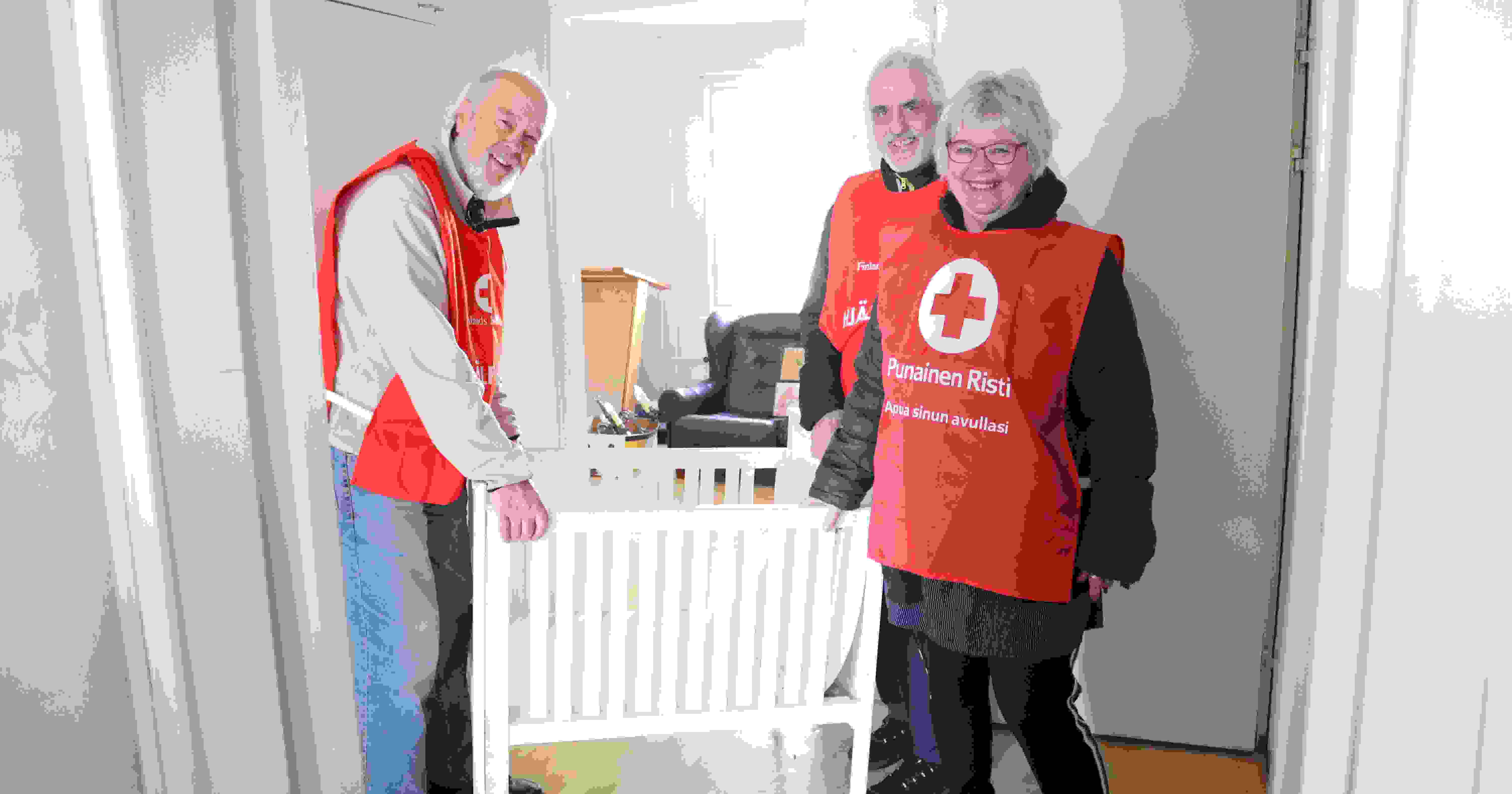 Tre leende personer i Röda Korsets frivilligvästar som står vid en vagga i en bostad som de inrett för flyktingar.