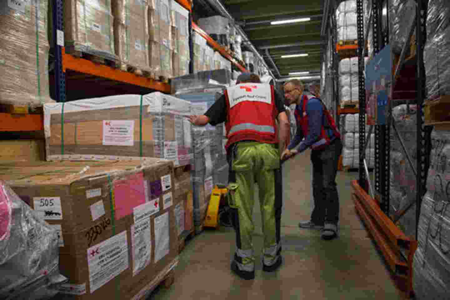 Två rödakorsanställda hämtar förnödenheter på logistikcentralen, där de höga och långa hyllorna är fulla av material.