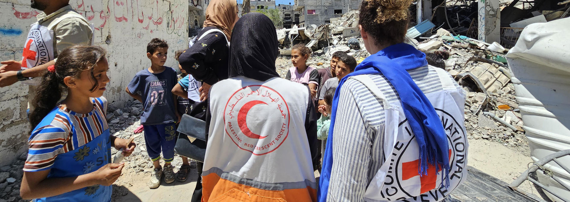 Anställda från Röda Korset och Röda Halvmånen talar med barn i ett förstört område i Gaza. I bakgrunden syns raserade byggnader.