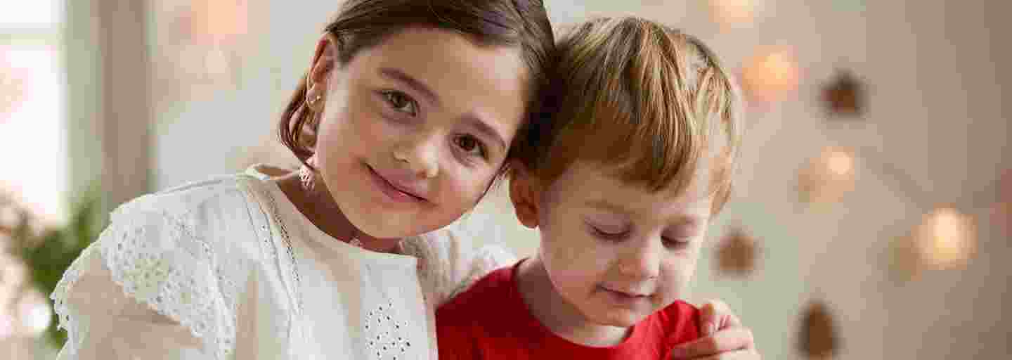 En leende flicka håller armen på sin brors axel i ett juldekorerat rum.