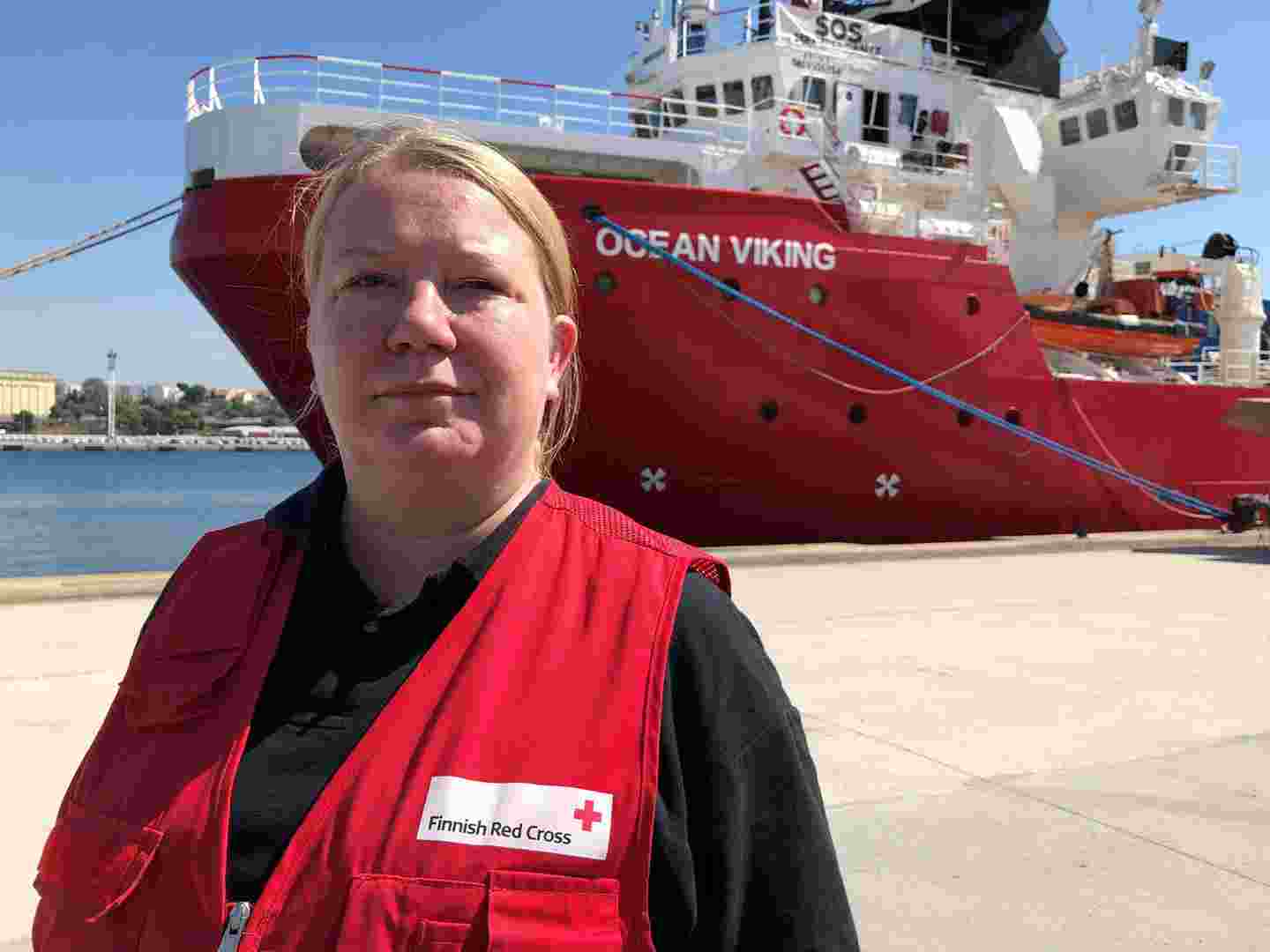 Biståndsarbetare klädd i Röda Korsets arbetsdräkt står framför ett fartyg på hamnområdet.