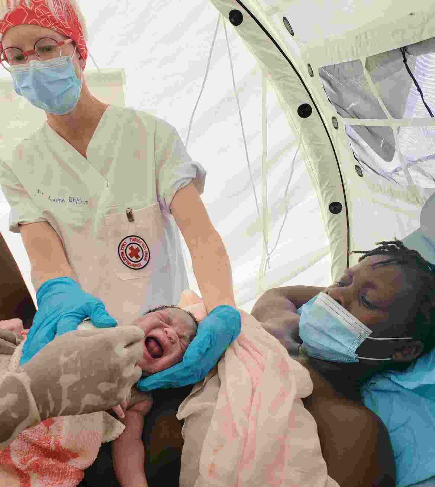 En läkare klädd i Röda Korsets kläder undersöker en nyfödd baby som är i moderns famn.