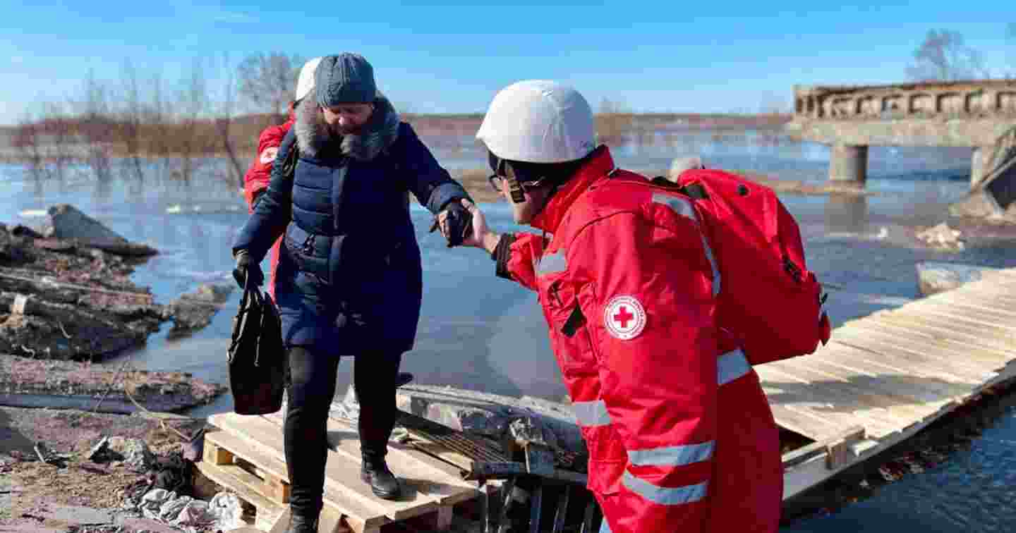 En frivillig på Röda Korset hjälper en äldre person att passera över en träbro.