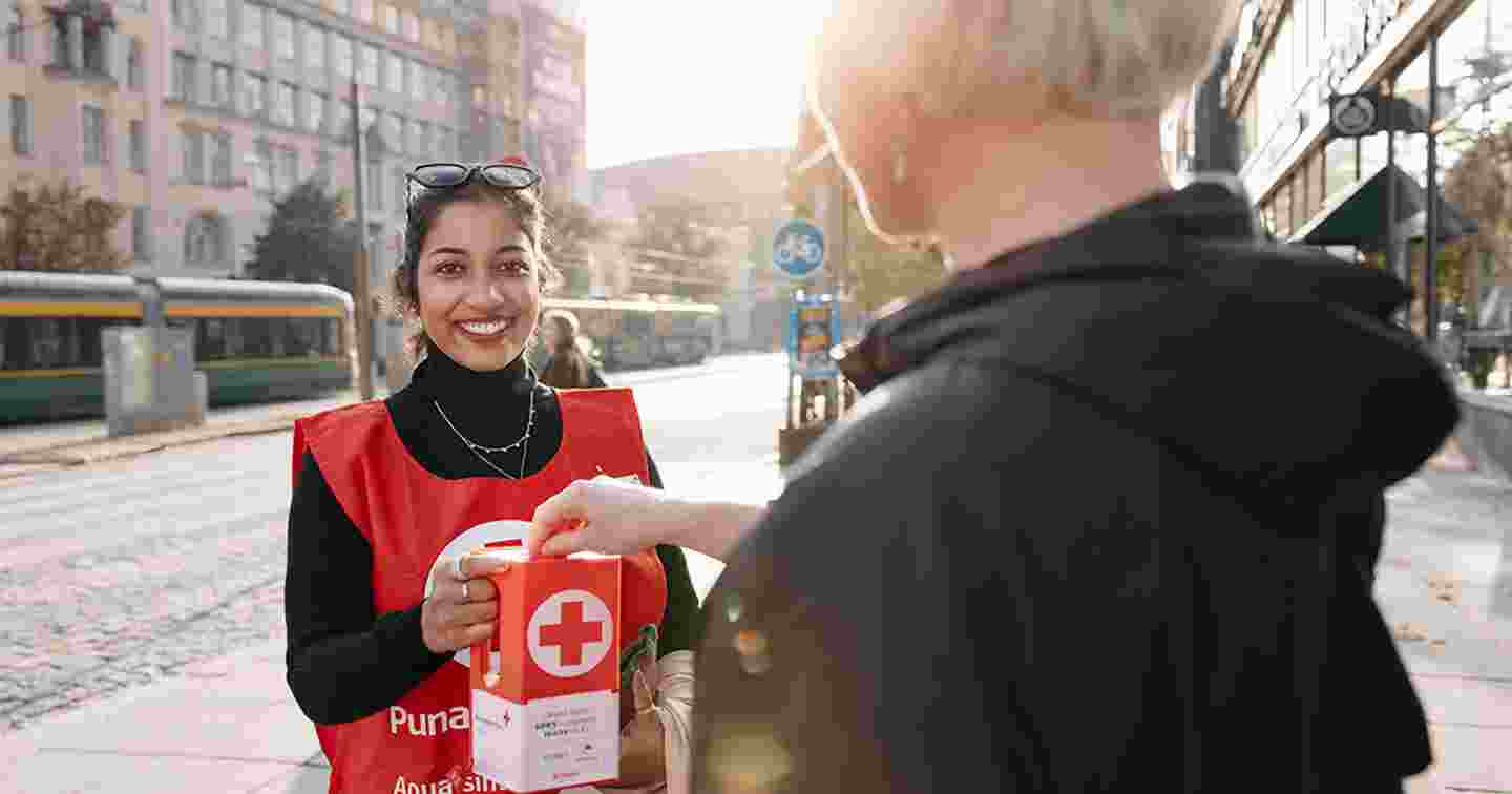En leende människa med Finlands Röda Korsets väst håller en insamlingslåda. En förbipasserande sätter en mynt i lådan.