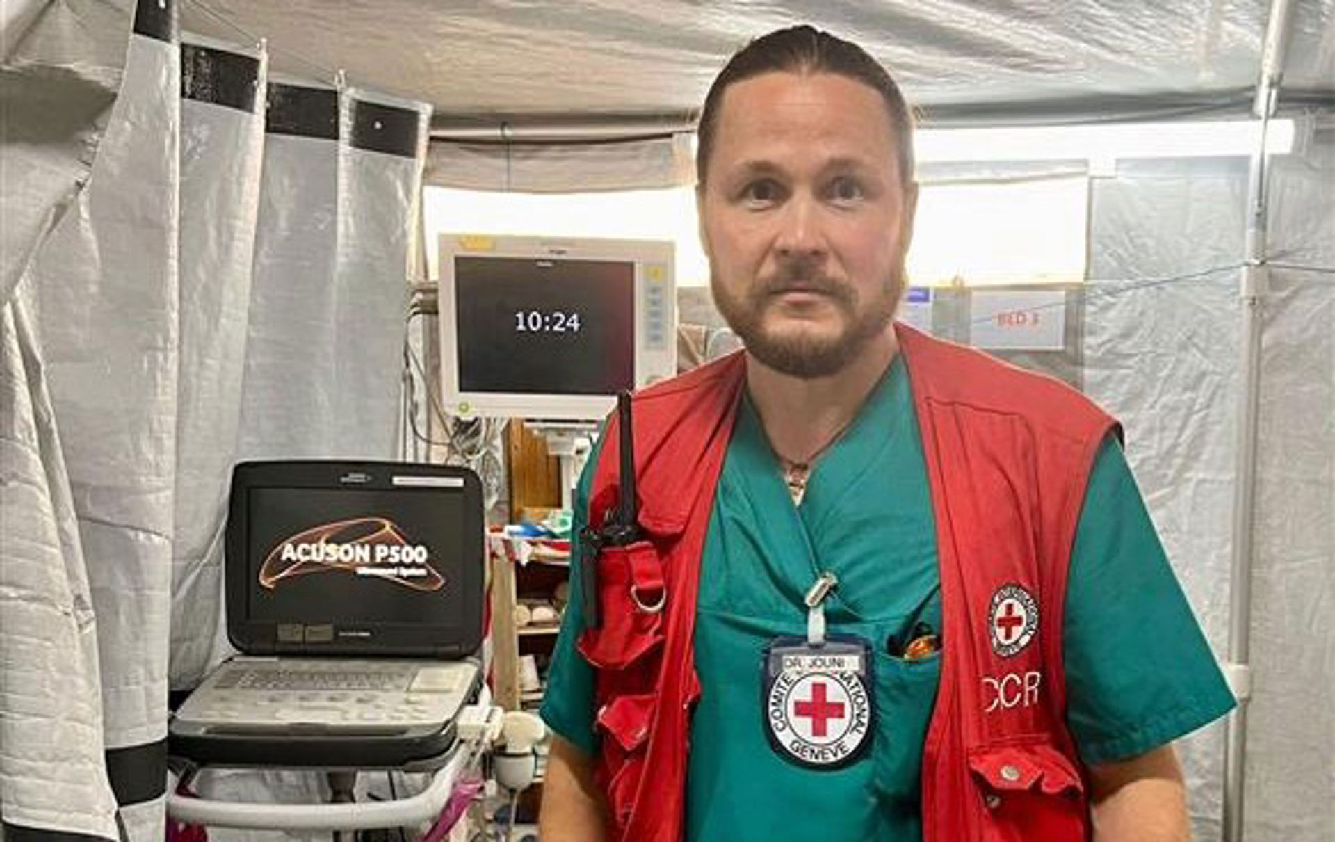 En person står i ett fältsjukhustält med sjukhusutrustning. Hen bär en grön sjukhusdräkt och en Röda Korsets väst med organisationens symbol samt ett ID-kort från Röda Korsets internationella kommitté. I sin bröstficka bär personen en radiotelefon.