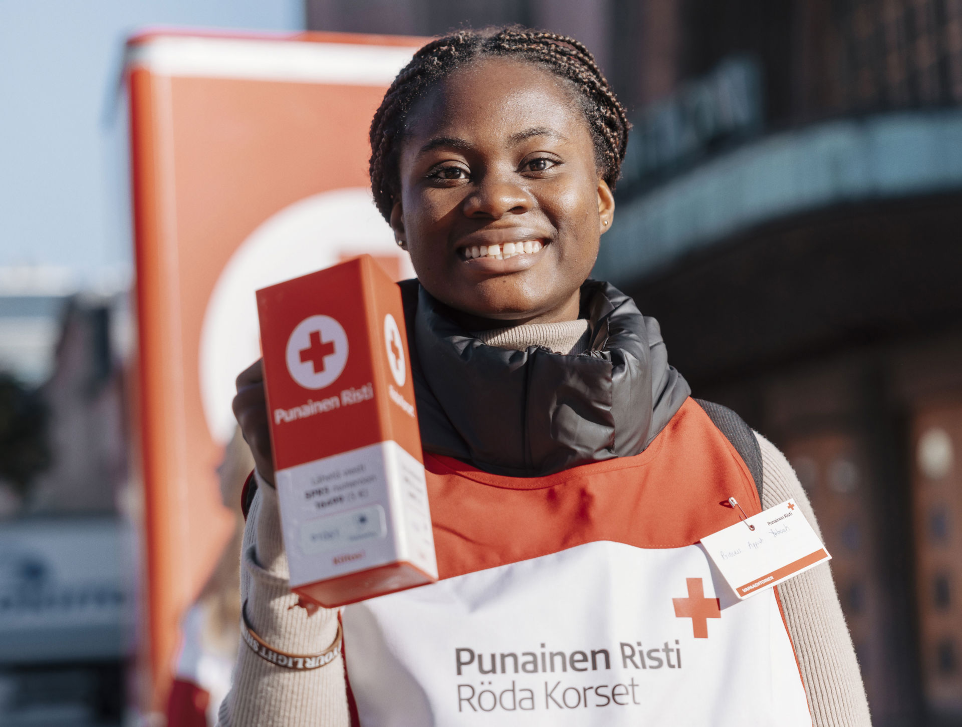 En leende frivillig håller i en insamlingsbössa i Hungerdagsinsamlingen. Personen har på sig en Röda Korsets frivilligväst med Röda Korsets logotyp och texten ”Punainen Risti Röda Korset”. I bakgrunden syns Röda Korsets stora insamlingsbössa och stadsmiljö.