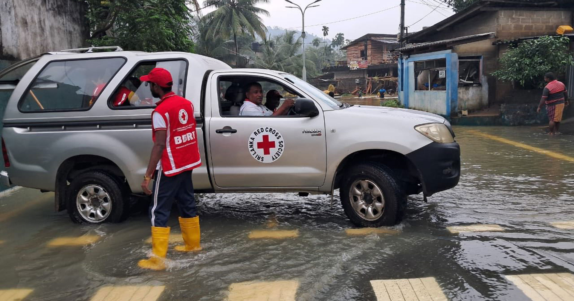 En hjälparbetare från Röda Korset står i översvämmat vatten med gula stövlar och röd väst, bredvid ett grått fordon med Röda Korsets logotyp på dörren. Fordonet befinner sig på en vattenfylld gata, och i bakgrunden syns tropiska träd, elledningar och byggnader i ett översvämningsområde.
