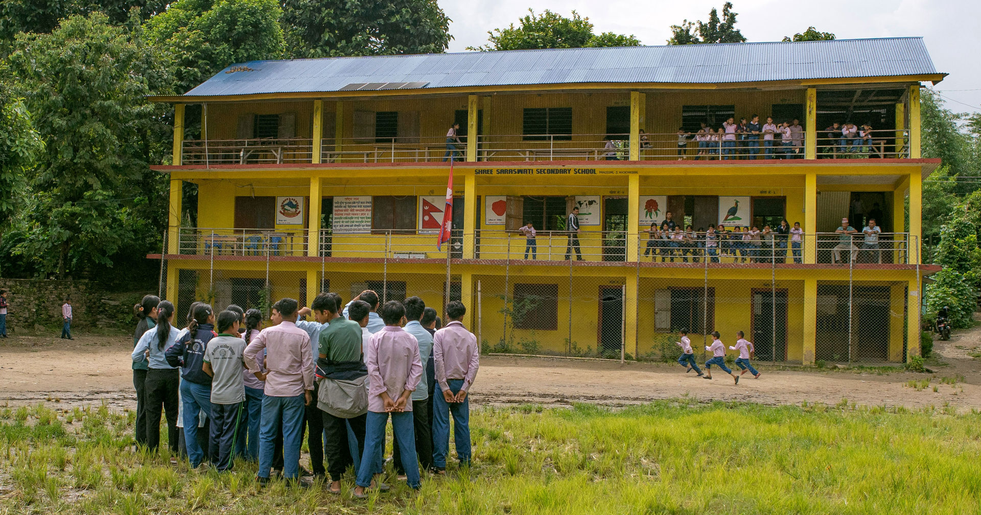 En grupp elever står på skolgården framför en gul skolbyggnad, medan en del av barnen springer på skolgården.