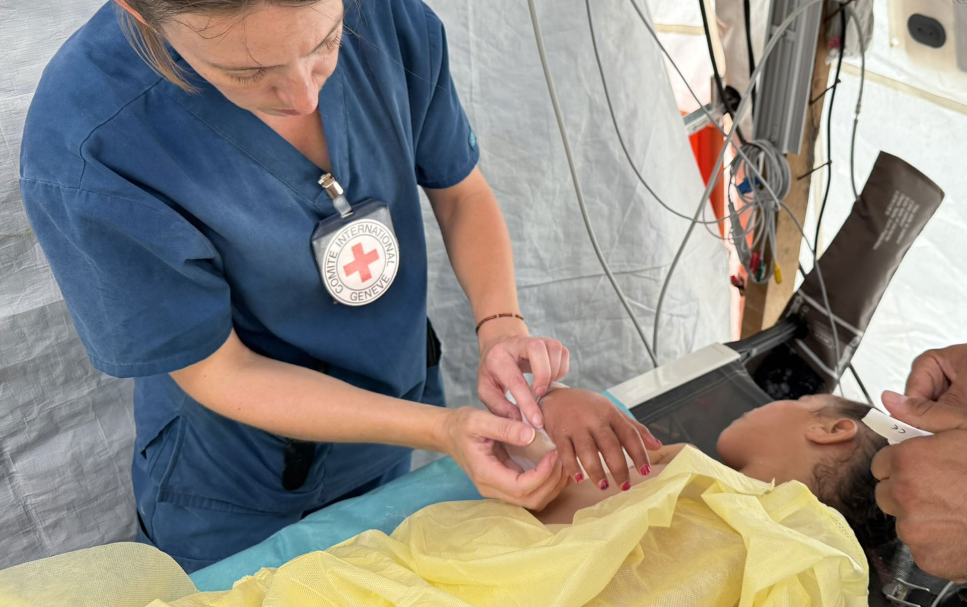 Röda Korsets fysioterapeut sätter gips på en barnpatients arm på fältsjukhuset. Patienten ligger i sängen med ett gult lakan över sig.