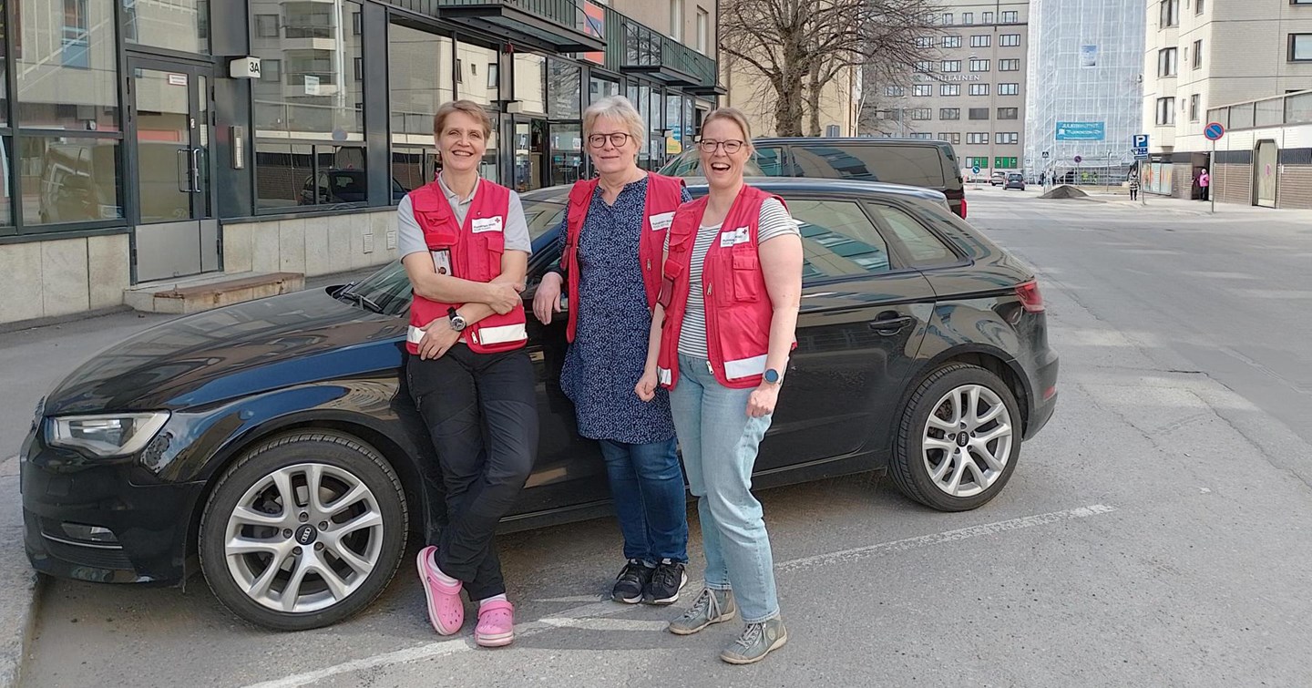 Tre leende personer i Röda Korsets kläder står framför bilen.