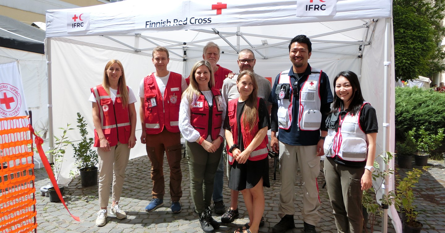Åtta biståndsarbetare i Röda Korsets arbetskläder står och ler framför ett tält som hör till hälsostationen.