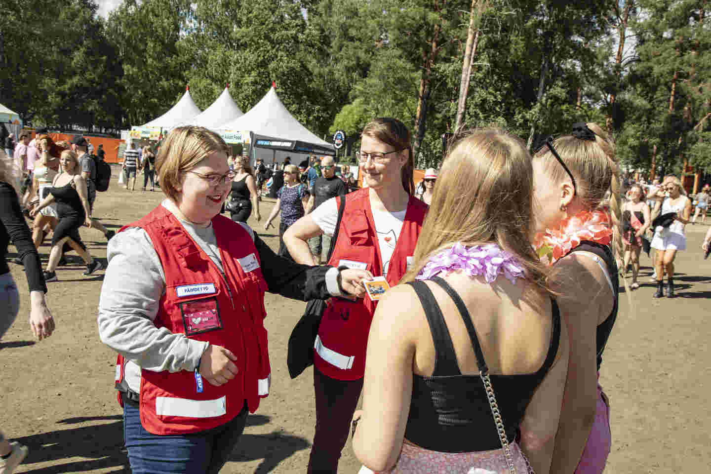 Röda Korsets frivilliga ger två personer en broschyr. I bakgrunden syns en solig festivalmiljö.