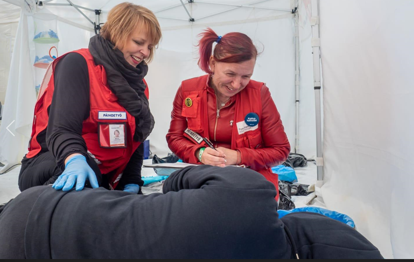 Två frivilliga vid Röda Korset ler mot en person som ligger på en fältsäng samt kontrollerar om personen mår bra.