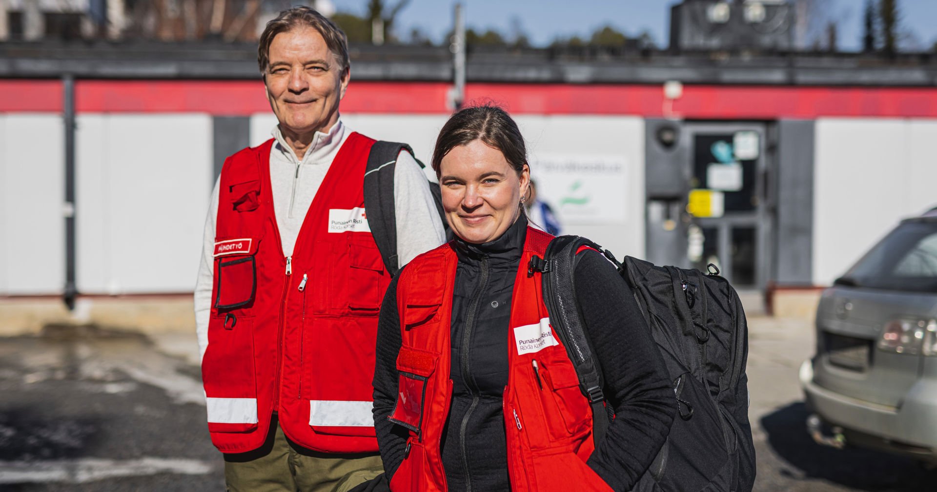 Kari är en äldre man som står med grått hår utomhus, leende och iklädd en röd Röda Korset-väst över en ljus dragkedjetröja.