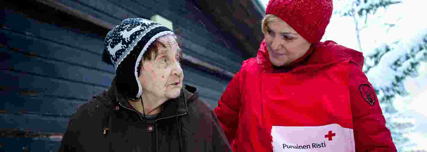 Två kvinnor står bredvid varandra i ett vintrigt landskap. Den yngre kvinnan i Röda Korsets väst för frivilliga tittar vänligt på den äldre.