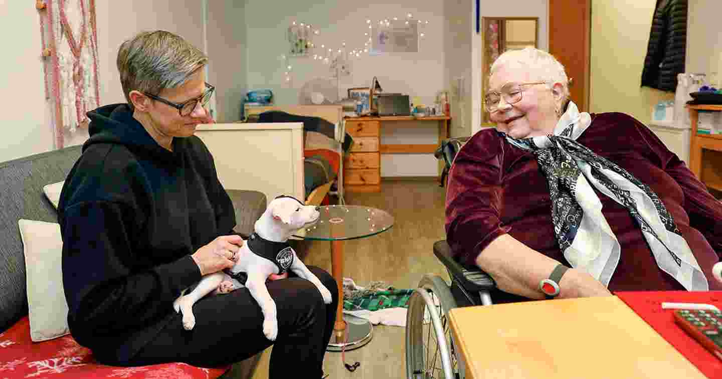 En frivillig och en äldre person sitter vid bordet och ler. Frivilligaren har en liten hund i famnen.