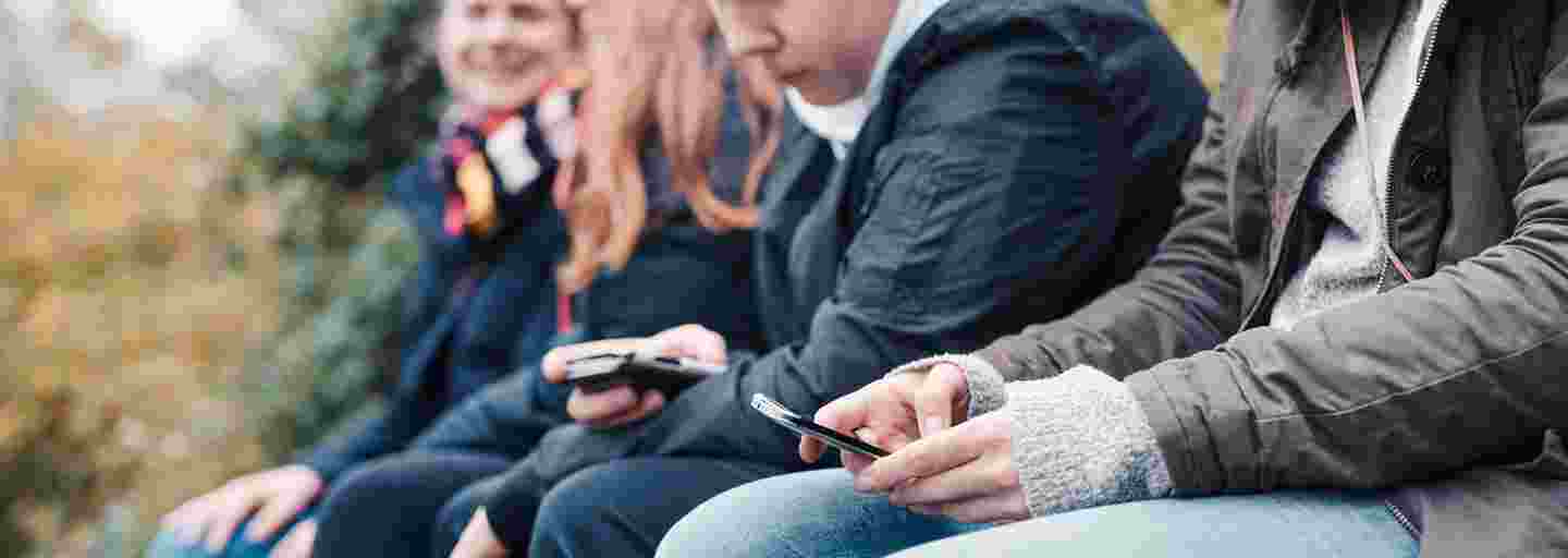 Fyra ungdomar sitter utomhus i rad med smarttelefoner i händerna.
