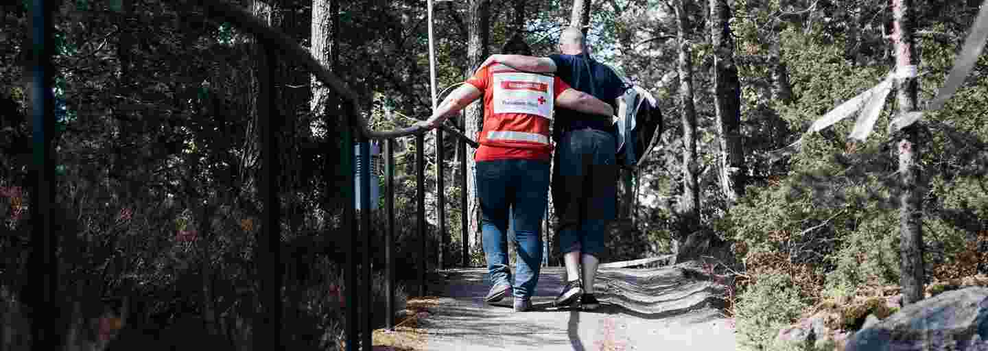 En man stöder sig på Röda Korsets frivillig när han går i uppförsbacke på en trädkantad väg.