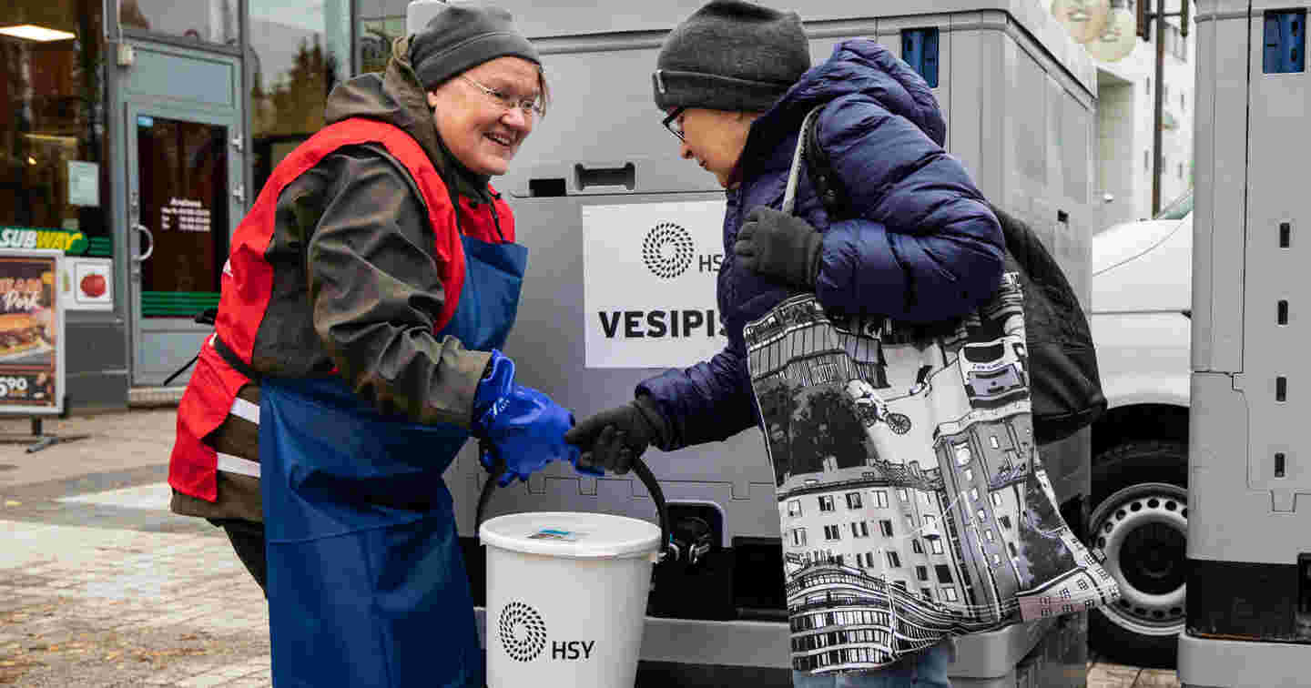 Hymyilevä nainen Punaisen Ristin vapaaehtoisliiveissä ojentaa kannellista ämpäriä naiselle, jonka vedenjakelu on katkennut. Punainen Risti suorittaa vedenjakelua kotimaan kriisitilanteissa.