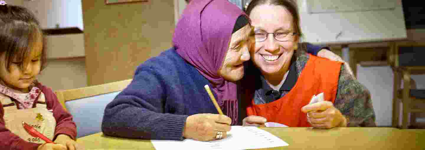 En kvinna i Röda Korsets frivilligväst lär en äldre invandrarkvinna och ett invandrarbarn. Den äldre kvinnan kramar den frivilliga.