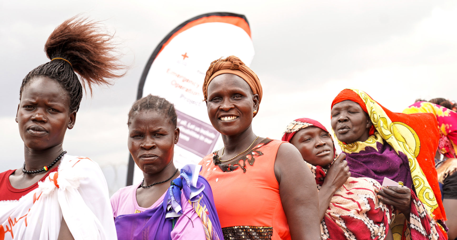 Människor står i kö ute i vinden med en banner bakom sig där Röda Korsets logotyp syns