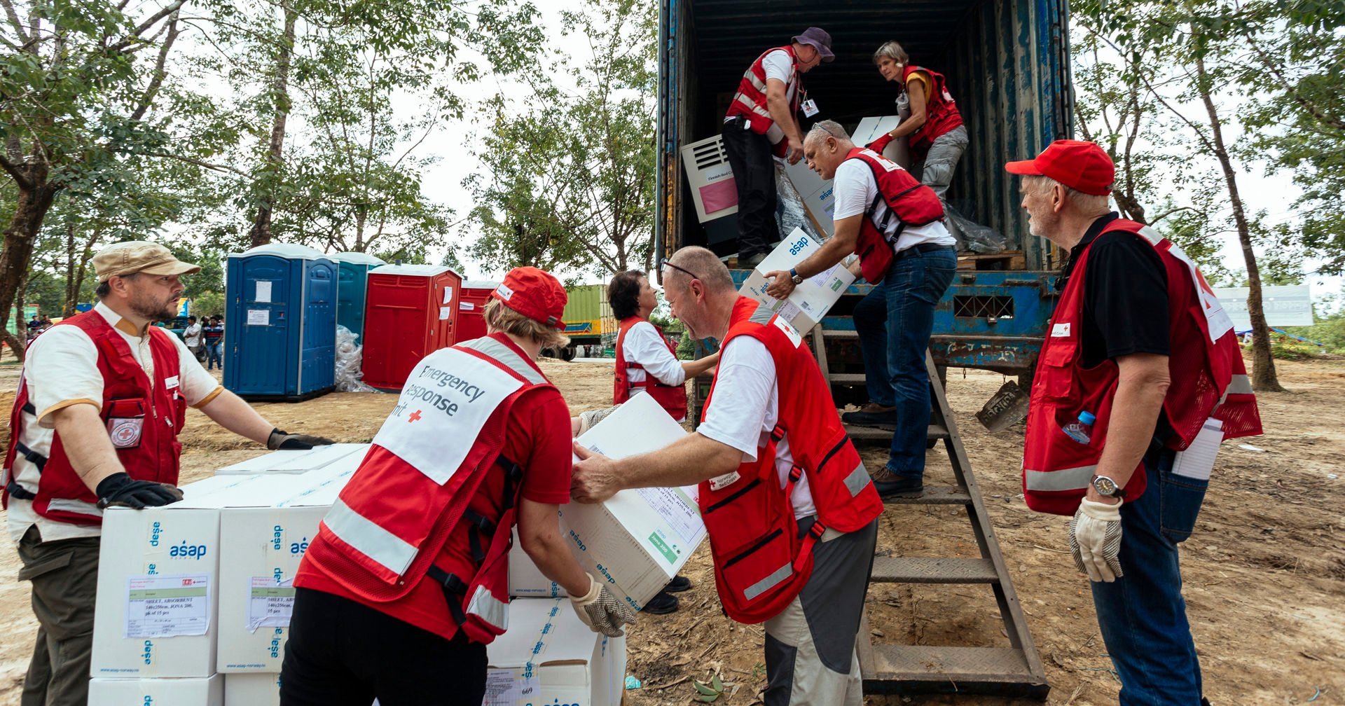 En grupp av Röda Korsets avustustyöntekijöitä lastar av avustustarvikelådor från en fraktcontainer och vidare ut på marken.