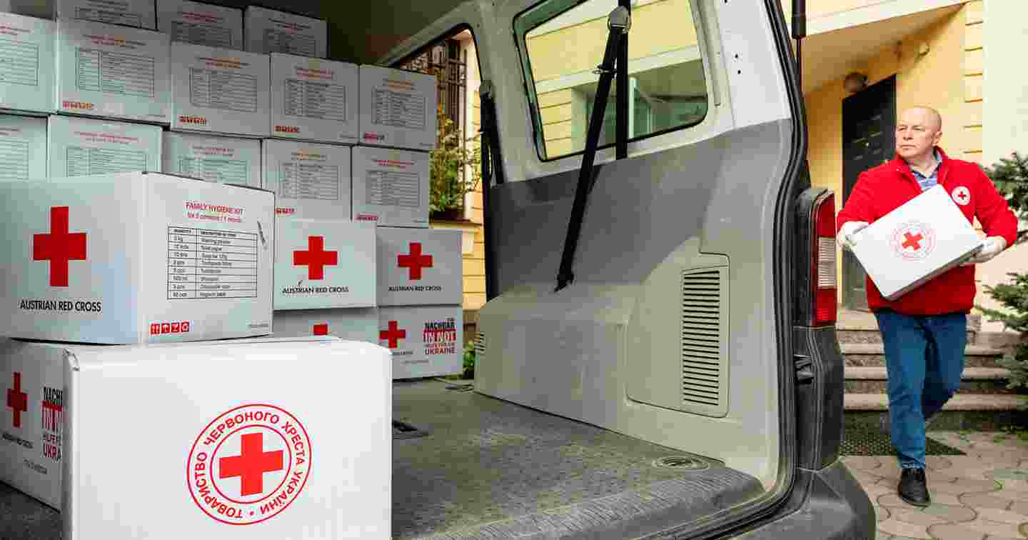 En person i Röda Korsets arbetskläder bär en låda med Röda Korsets biståndsmaterial till en paketbil, som är halvfull med biståndsmaterial.