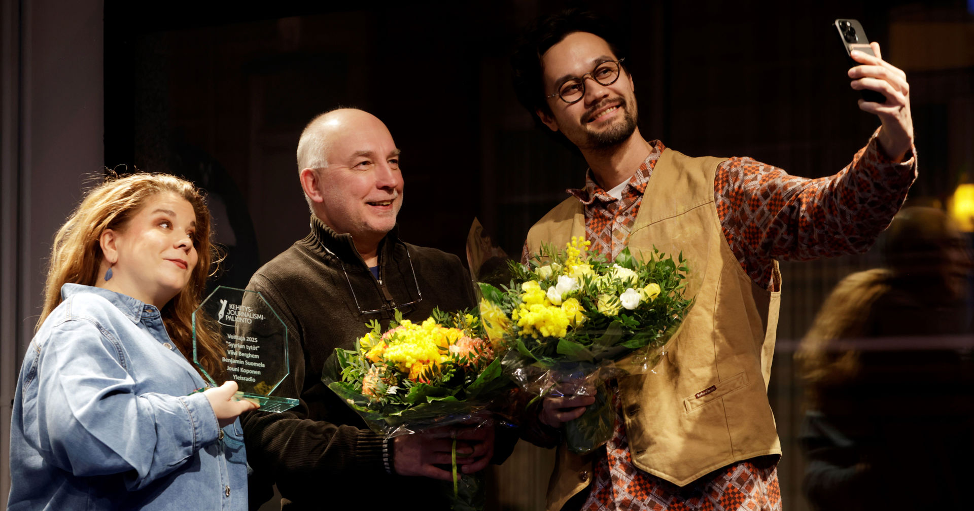 Tre personer står bredvid varandra inomhus och håller i blombuketter och ett pris, samtidigt som en av dem tar en gruppselfie.