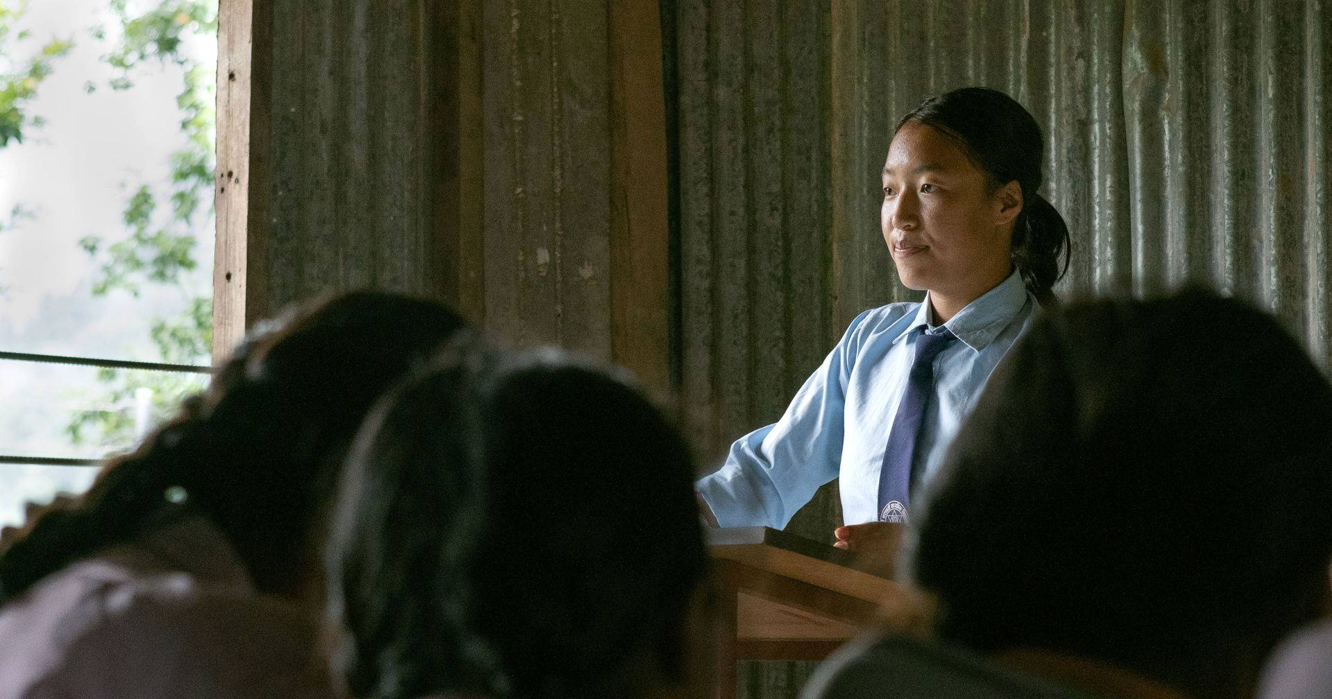 En ung person står i ett klassrum bakom en kateder och bär skoluniform. I förgrunden finns elever som lyssnar, med klassrummets väggstrukturer och fönster i bakgrunden.