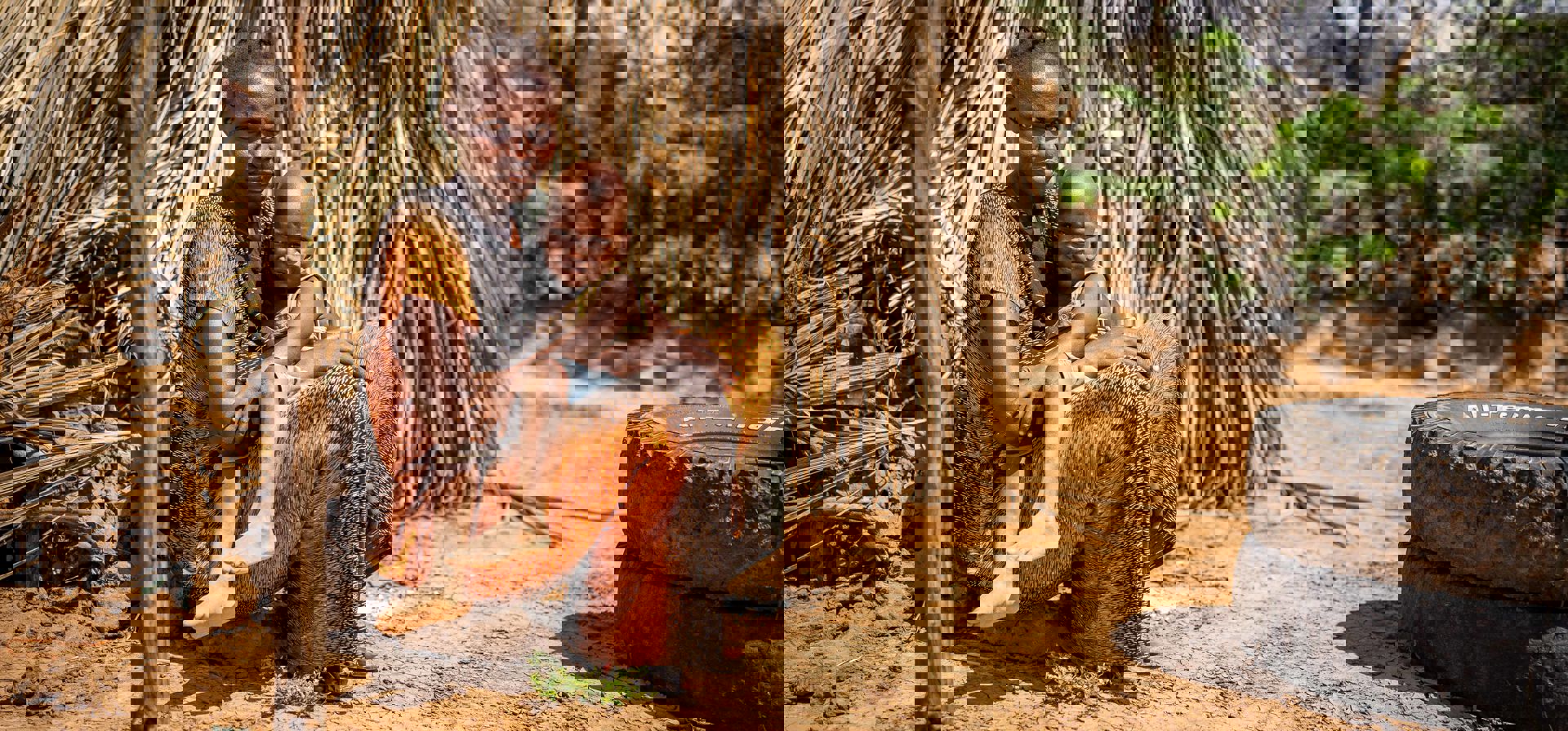 En kvinna sitter med sitt barn i famnen bakom en byggnad av lera och löv.