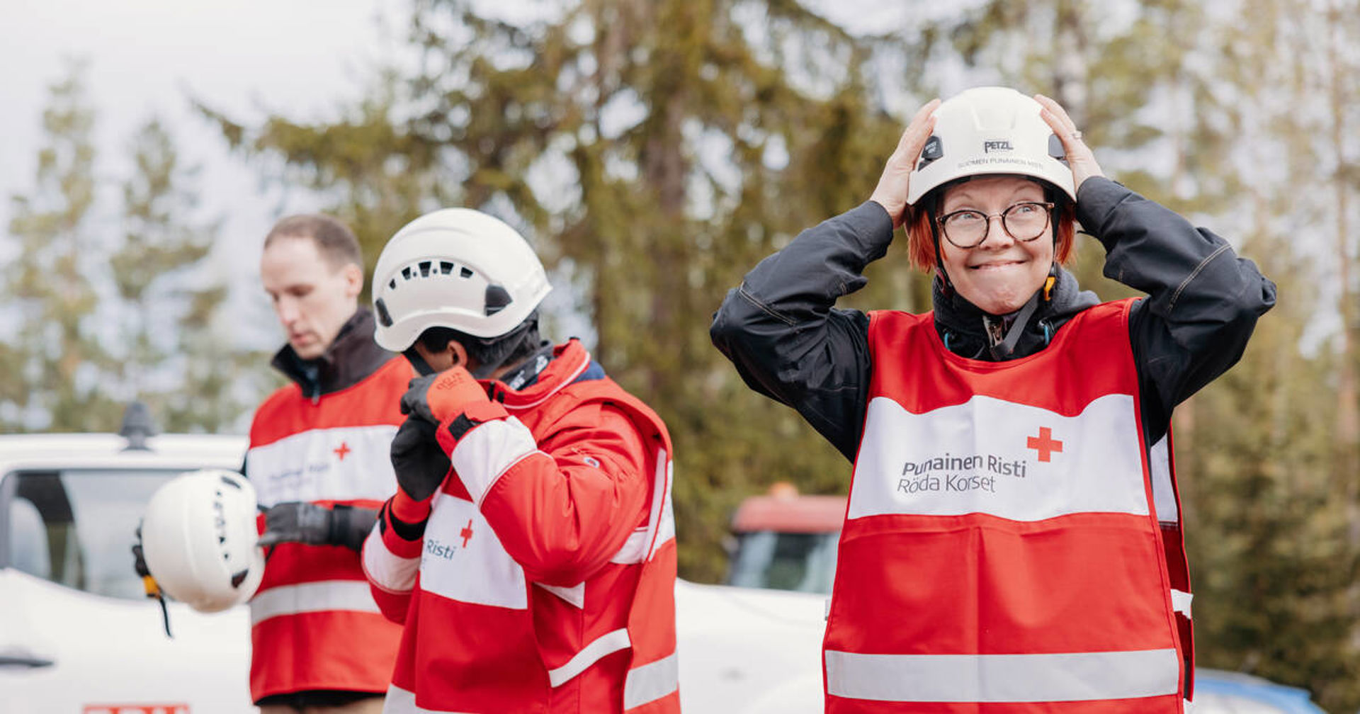 Tre personer i Röda Korsets röda skyddsvästar står utomhus vid ett fordon i en skogsmiljö. Den främsta personen tar på sig en vit skyddshjälm, medan två personer i bakgrunden också förbereder sig och hanterar hjälmar och utrustning. På västarna står “Punainen Risti / Röda Korset”.