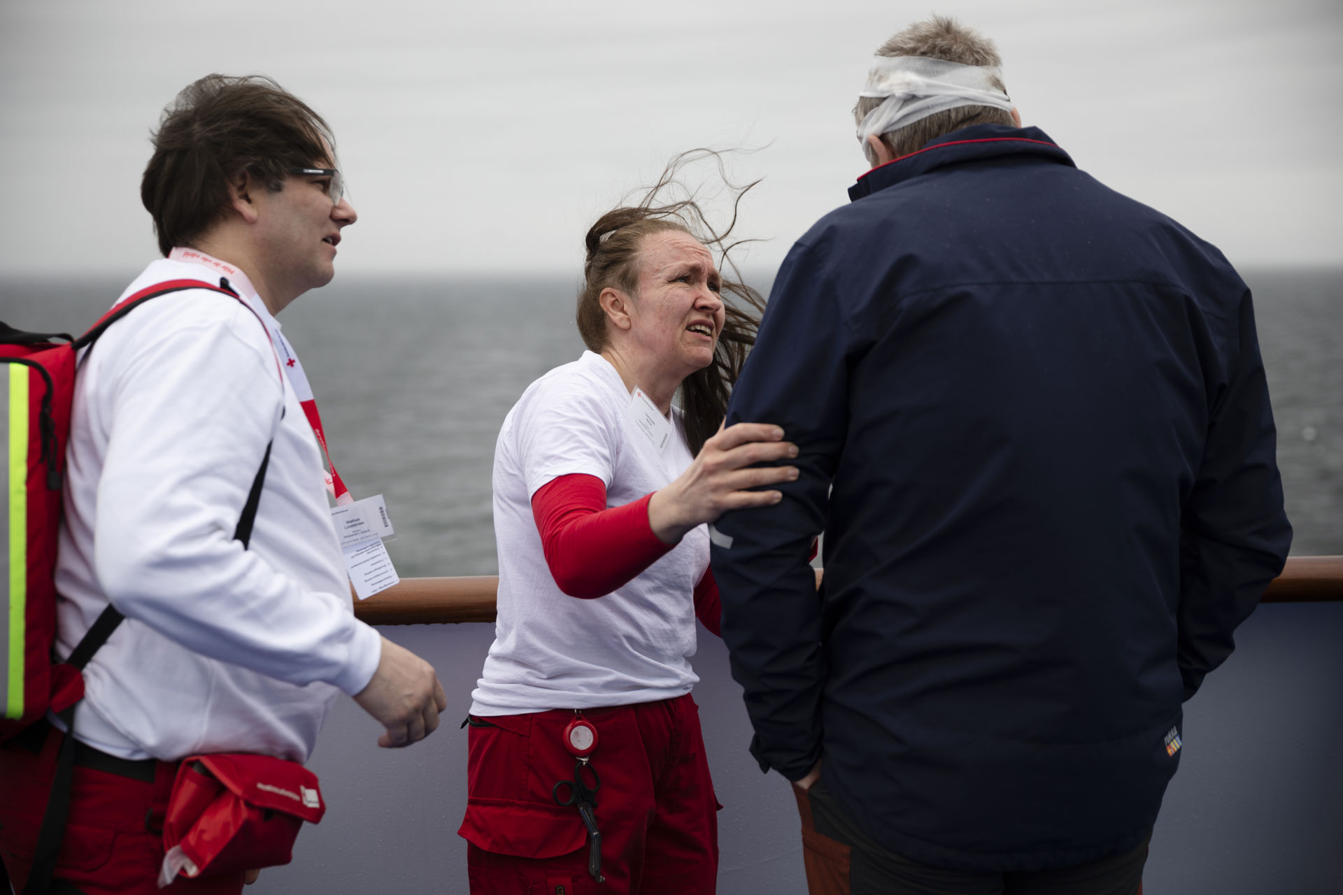 Två frivilliga från svenska Röda Korset talar med en målperson, som har ett bandage runt huvudet. Östersjön syns i bakgrunden. Fotot har tagits från fartygets däck.