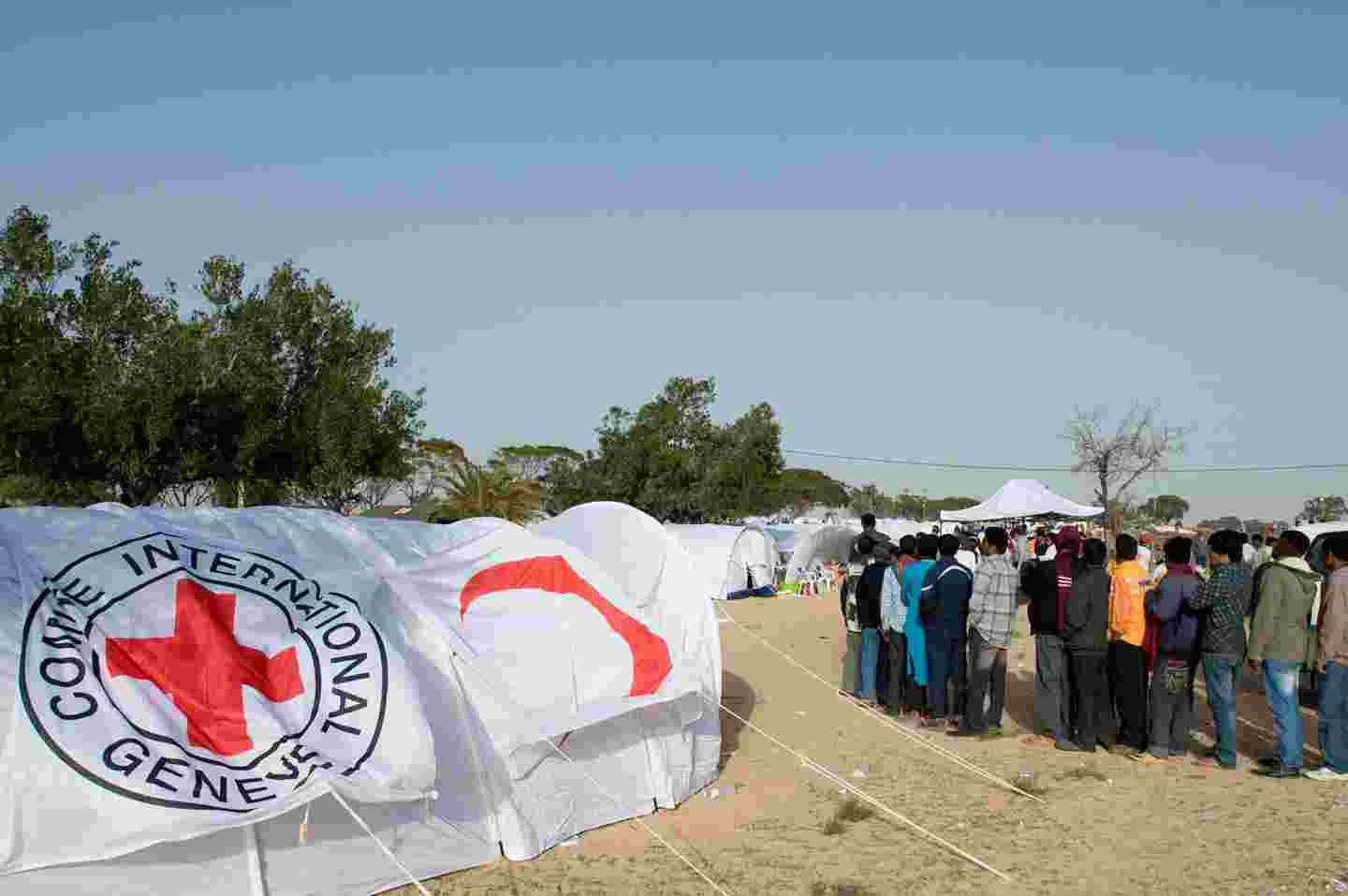Människor står i kö på flyktingläger mitt bland tält som är försedda med Röda Korsets kännetecken.