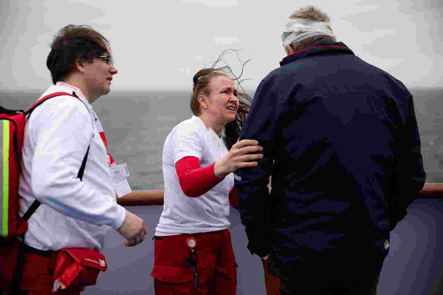 Två frivilliga från svenska Röda Korset talar med en målperson, som har ett bandage runt huvudet. Östersjön syns i bakgrunden. Fotot har tagits från fartygets däck.