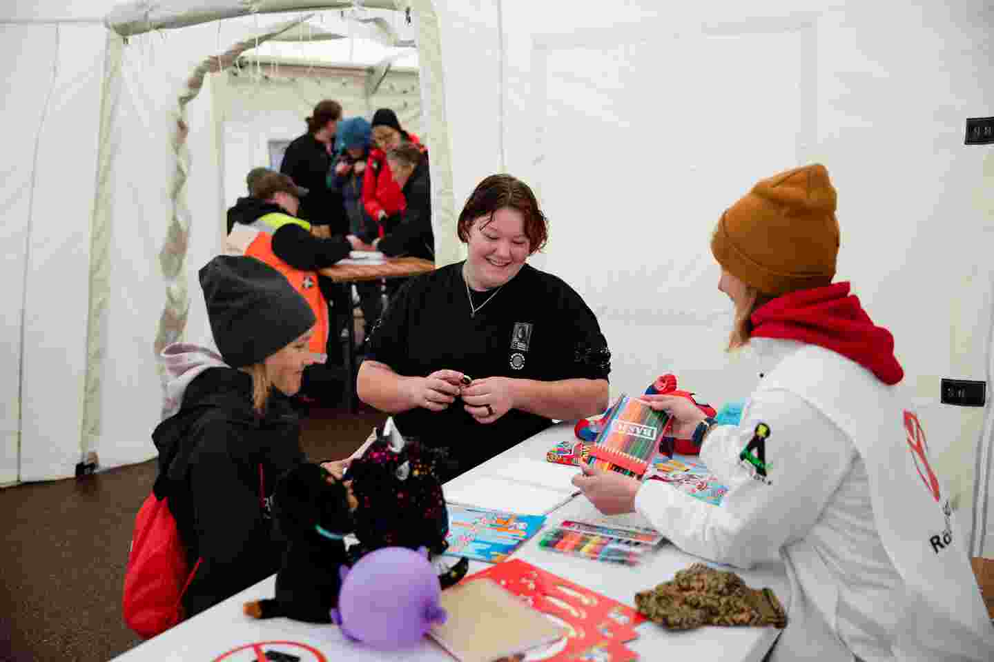 En frivillig från Rädda Barnen visar färgpennor för ett barn i tältet. Barnets mor tittar leende på bredvid.