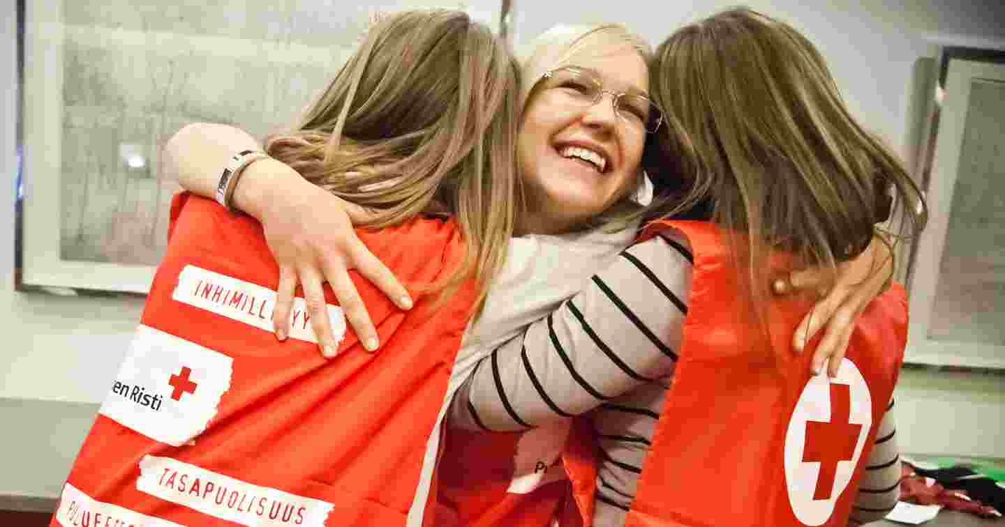 Tre unga kvinnor i Röda Korsets västar för frivilliga ler och kramar om varandra.