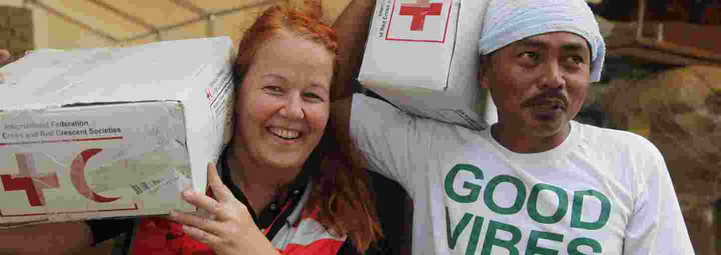 En av Röda Korsets biståndsarbetare och en lokal hjälpare bär biståndsmaterial med ett leende på läpparna.
